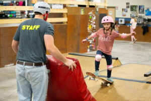 A young skateboarder is about to drop in on a small ramp while an instructor holds a crash pad for her.