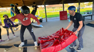 A skate coach holds a crash pad for a young rider about to roll in on a mobile skate ramp
