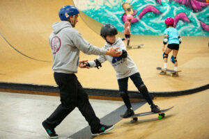 A skate coach helps a young skater learn to go up and down a ramp