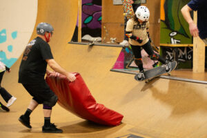 A skate coach holds a crash pad for a rider learning a new trick in the mini ramp.